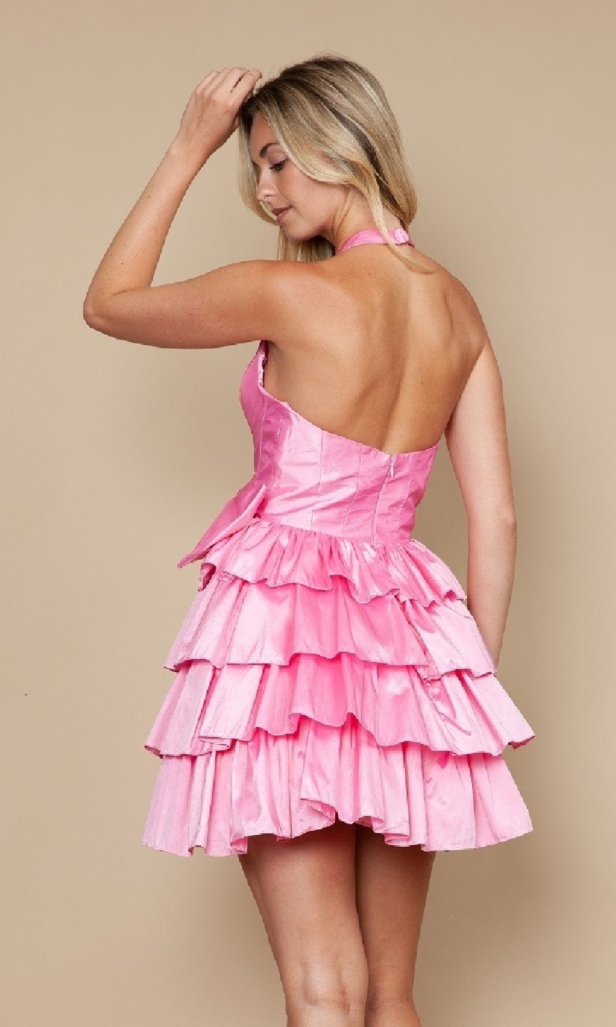 A person poses with one arm raised against a beige background, wearing the Ruffled Short Halter Hoco Dress Poly USA 9796— a sleeveless, open-back party dress in pink tiered ruffles.